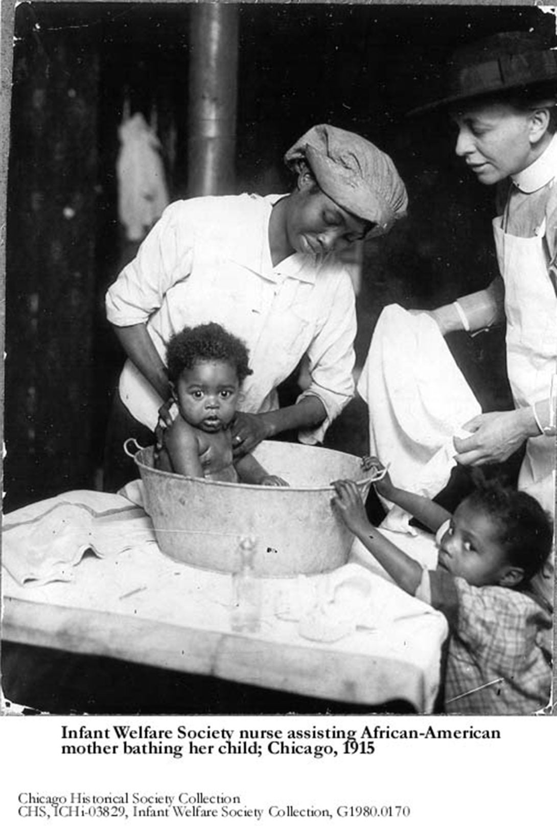 historical photo of a white nurse assisting an African-American woman bathing her baby