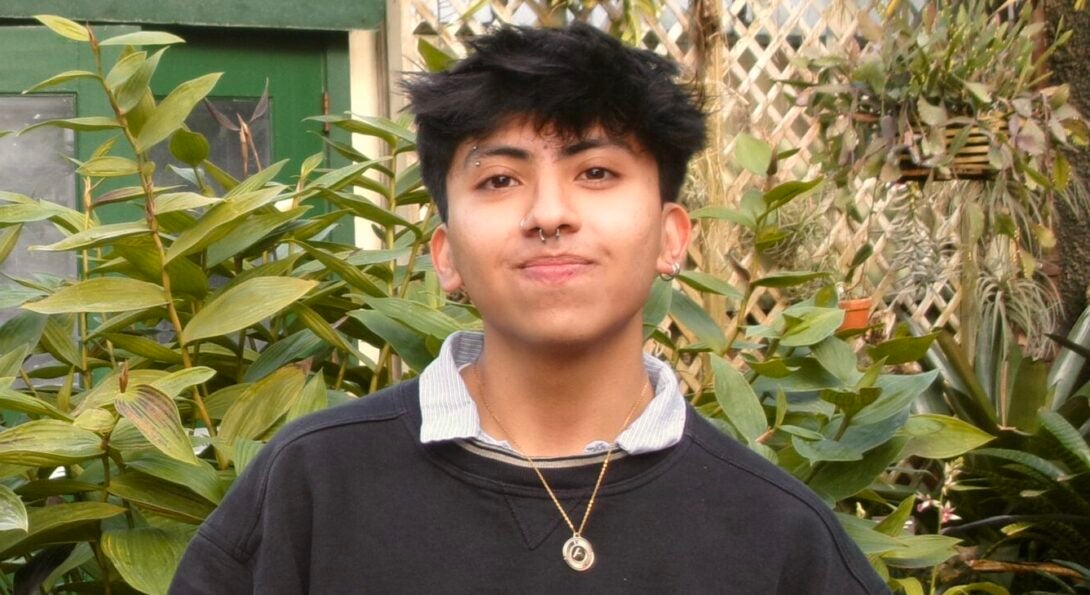 Rodrigo Vega smiles at the camera in a dense garden. Rodrigo has bronze skin and short black hair and is wearing a nice black shirt and a pendant necklace.