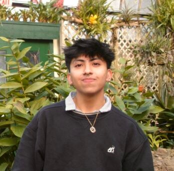 Rodrigo Vega smiles at the camera in a dense garden. Rodrigo has bronze skin and short black hair and is wearing a nice black shirt and a pendant necklace.
                  