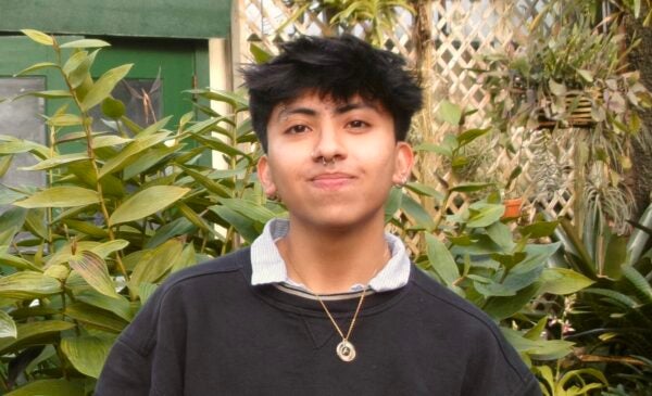 Rodrigo Vega smiles at the camera in a dense garden. Rodrigo has bronze skin and short black hair and is wearing a nice black shirt and a pendant necklace.